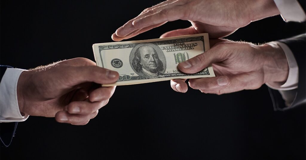 Two men in suits hold a dollar bill, symbolizing financial dealings related to conspiracy charges.